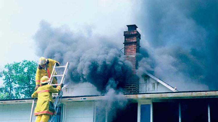 Fire fighters at the top of a ladder preparing to put out a house fire with dark smoke exiting the roof.